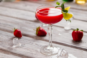 Red fruit cocktail with mint and lemon garnish on a wooden deck with strawberries scattered in the background