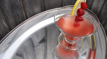 Rosy pink cocktail with raspberries and lemon garnish on a silver tray with a weathered gray fence in the background